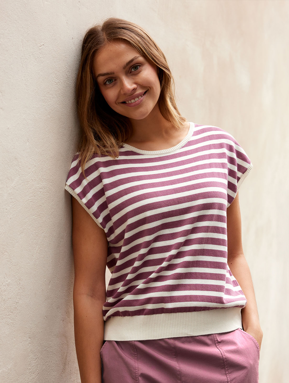 Frau trägt weiß violett gestreiftes Shirt mit violetter Hose und lehnt an einer Wand