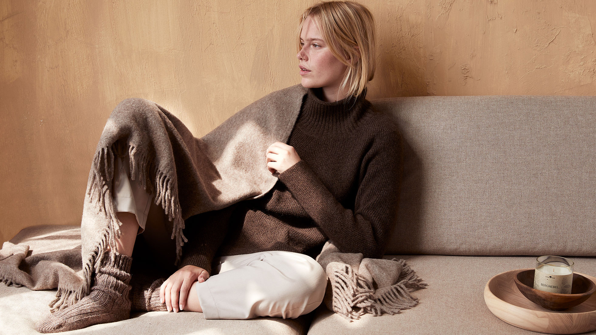 Frau in brauner Loungwear mit Wohndecke auf einem braunen Sofa