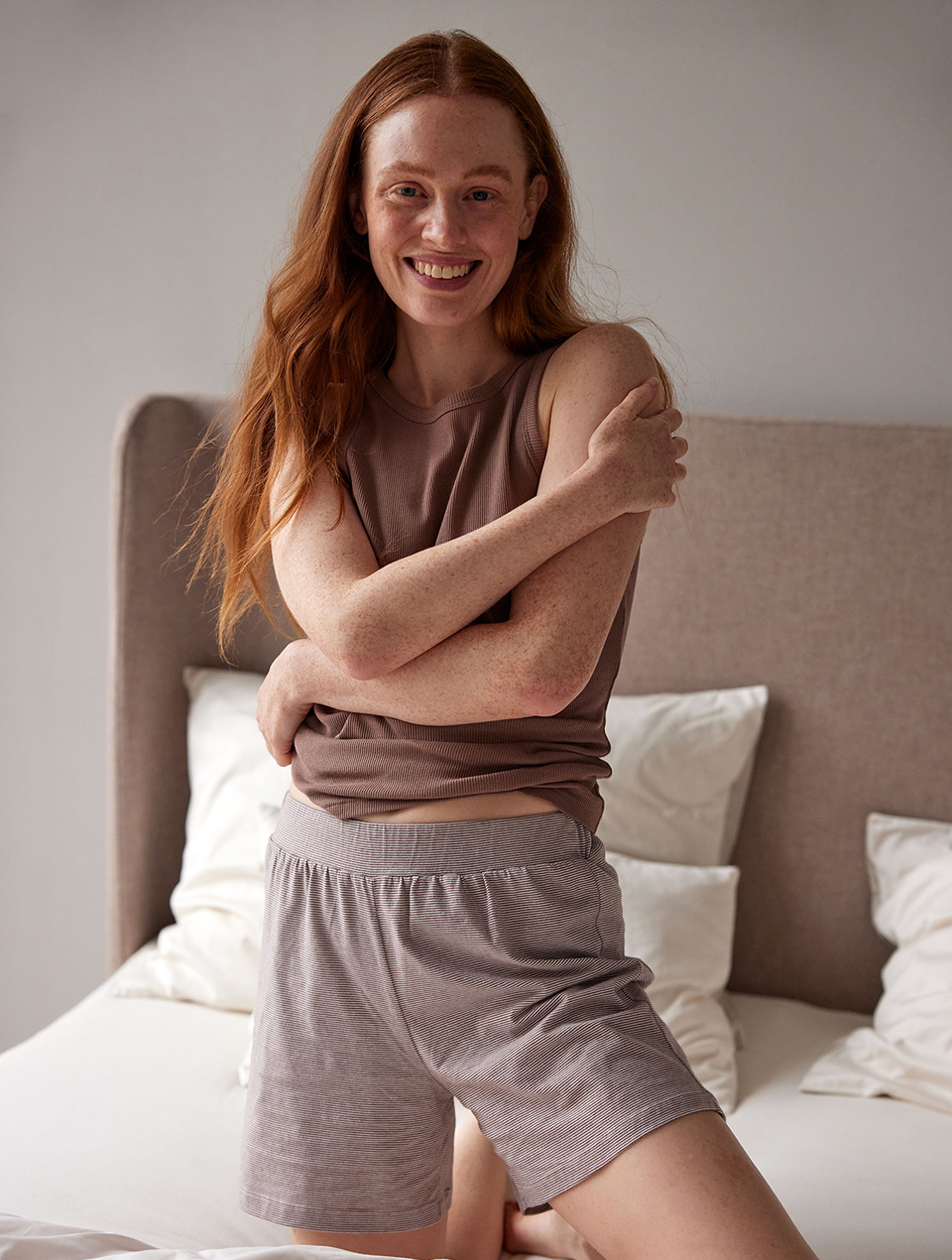 Frau trägt braunes Schlaftop und Shorts und kniet auf einem Bett