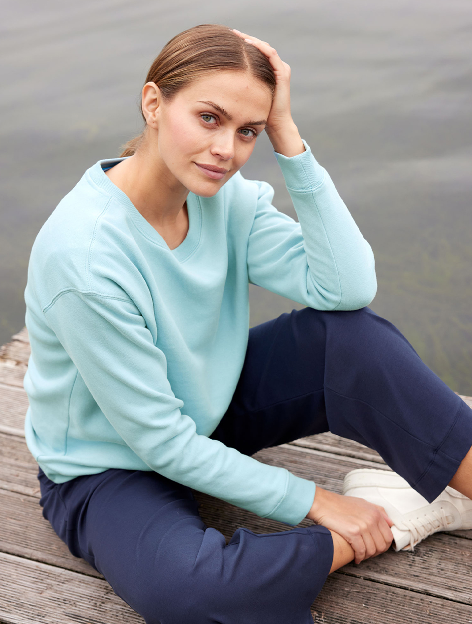 Frau trägt hellblaues Sweatshirt und dunkle Hose und sitzt am Wasser