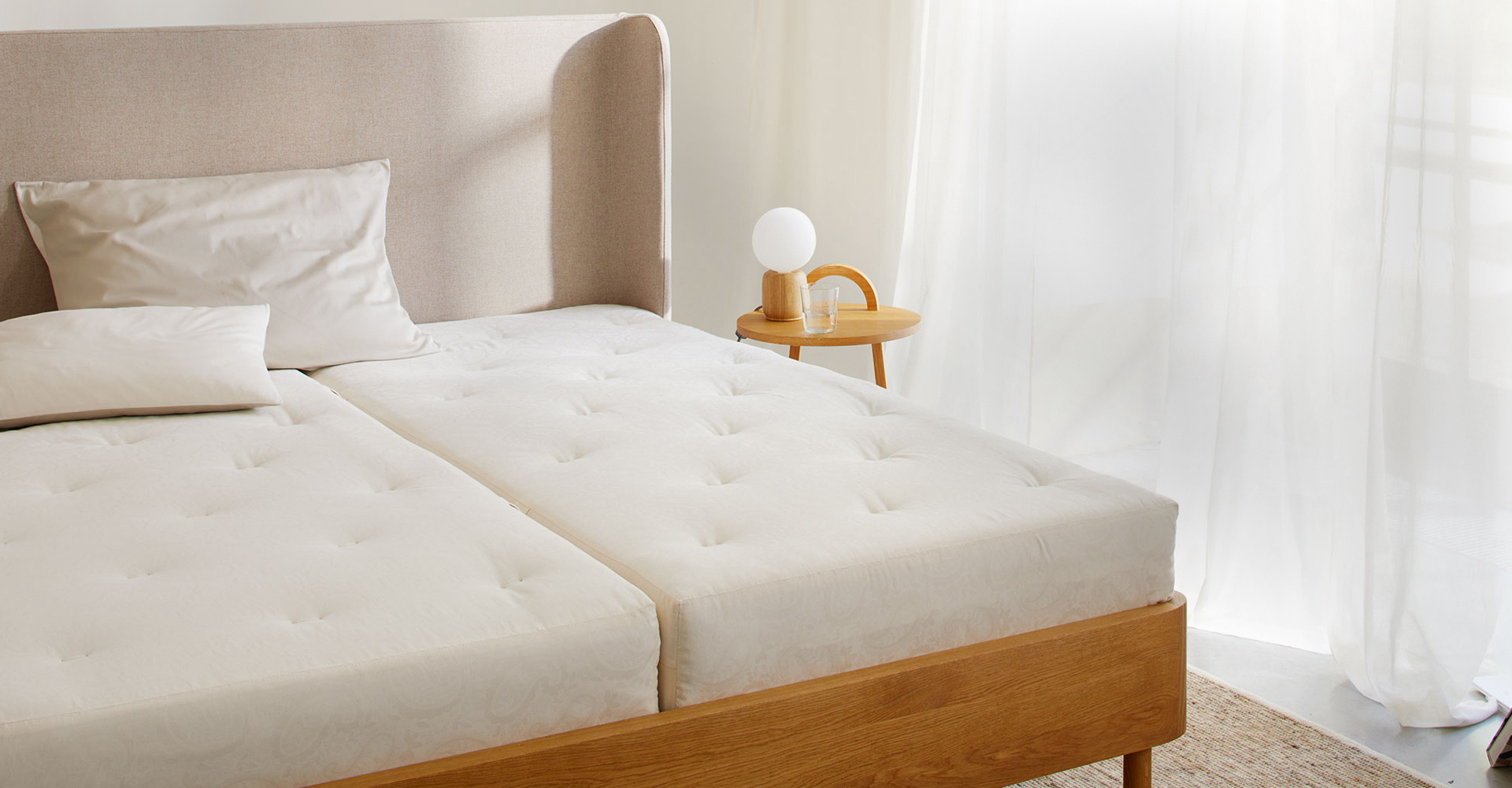 zwei Naturmatratzen in einem Massivholzbett mit Betthaupt vor einem großen Fenster mit weißen Vorhängen