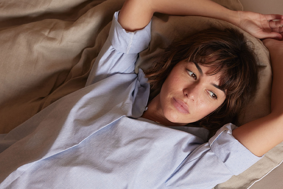 Eine dunkelhaarige Frau streckt die Arme über den Kopf und liegt auf einer grauen Bettwäsche