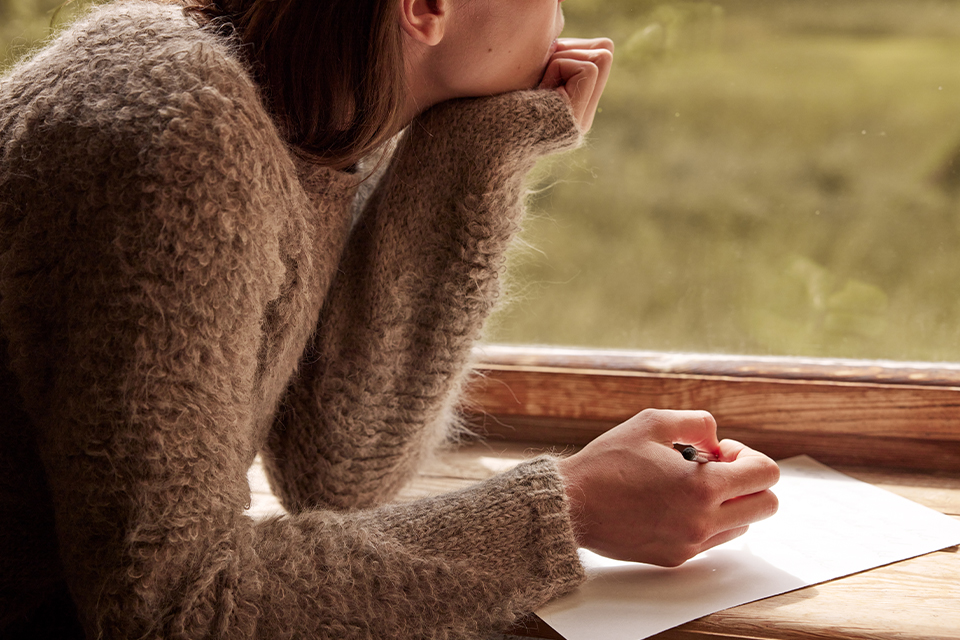 Frau schreibt auf ein Blatt Papier und blickt dabei nachdenklich aus dem Fenster