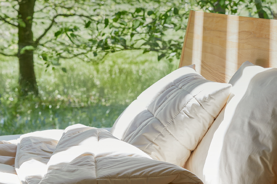 Massivholzbett mit Schlafdecken und Kopfkissen vor einer Wiese mit Bäumen