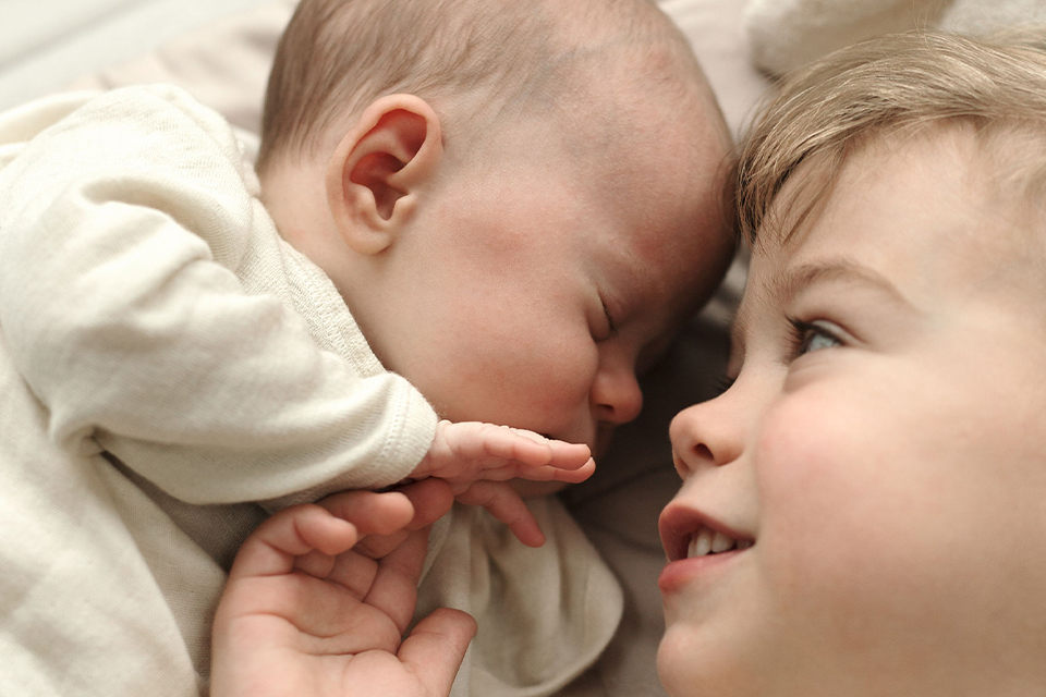 Kleinkind blickt liebevoll auf schlafendes Baby und hält sanft dessen Hand
