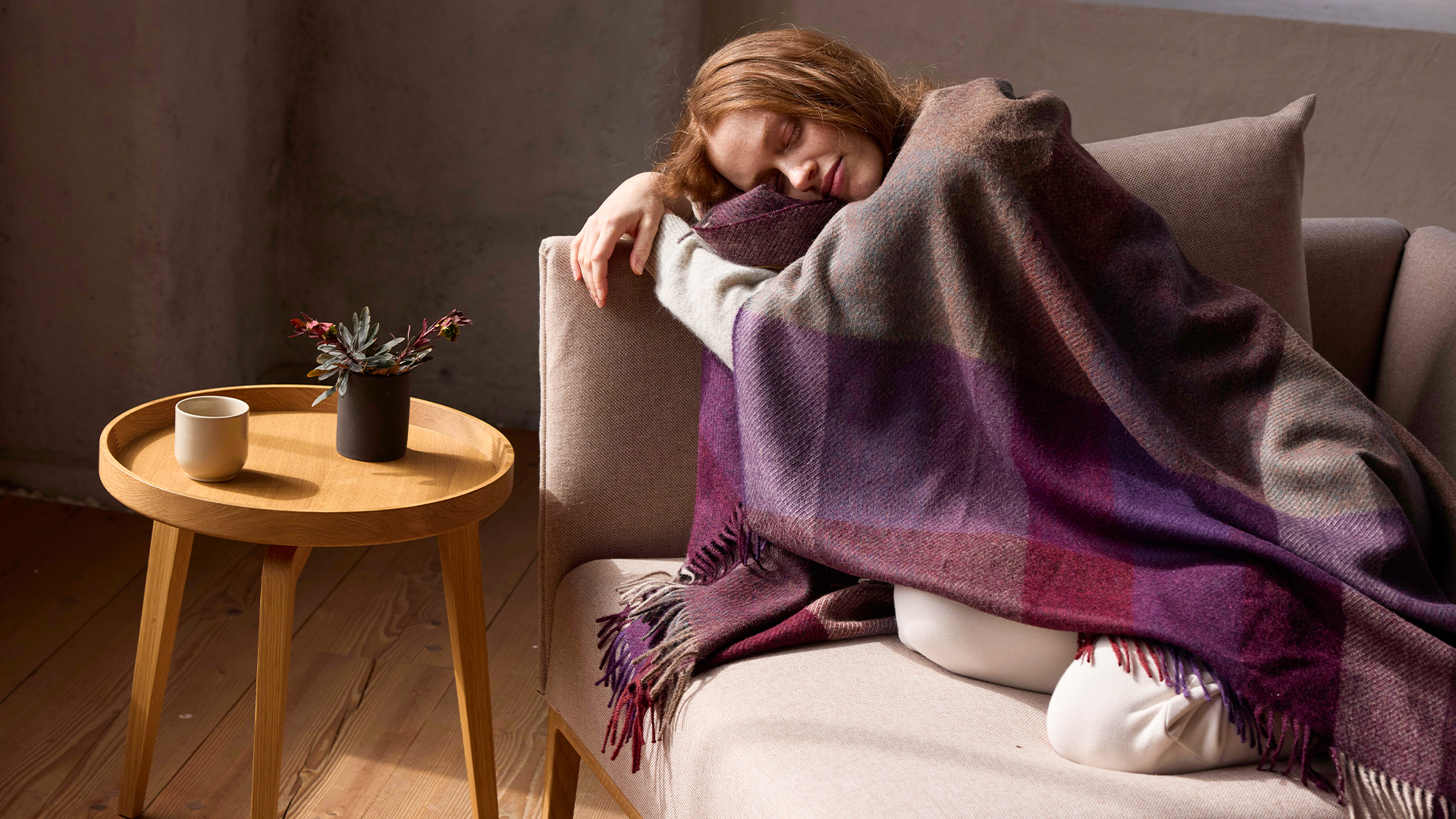Frau auf Sofa mit Decke neben kleinem Tisch mit Tasse