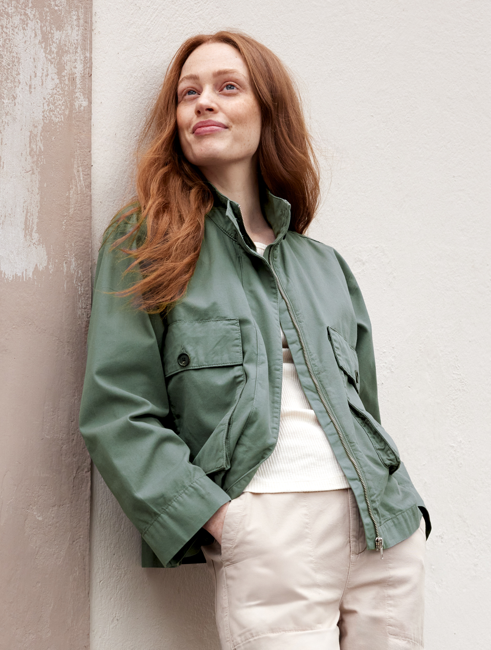 Frau mit grüner Übergangsjacke und beiger Hose lehnt an einer Wand