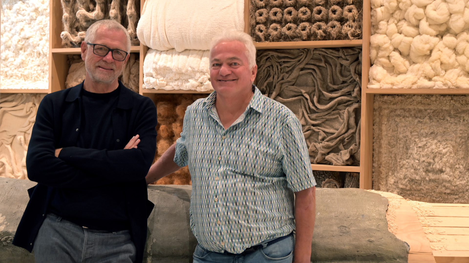 Zwei Männer stehen lachend vor einer Wand aus Naturmaterialien