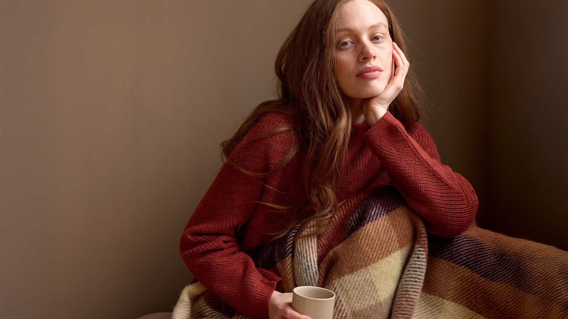 Frau in eine Wohndecke gehüllt und einer Tasse in der Hand auf einem Sofa sitzend