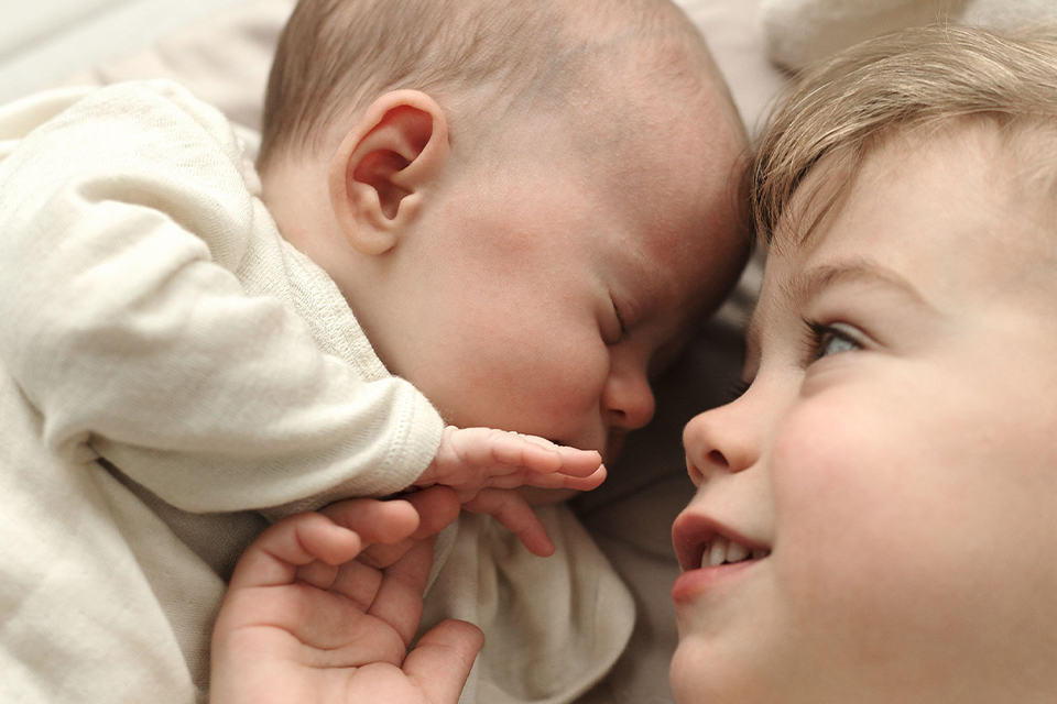 Kleinkind blickt liebevoll auf schlafendes Baby und hält sanft dessen Hand