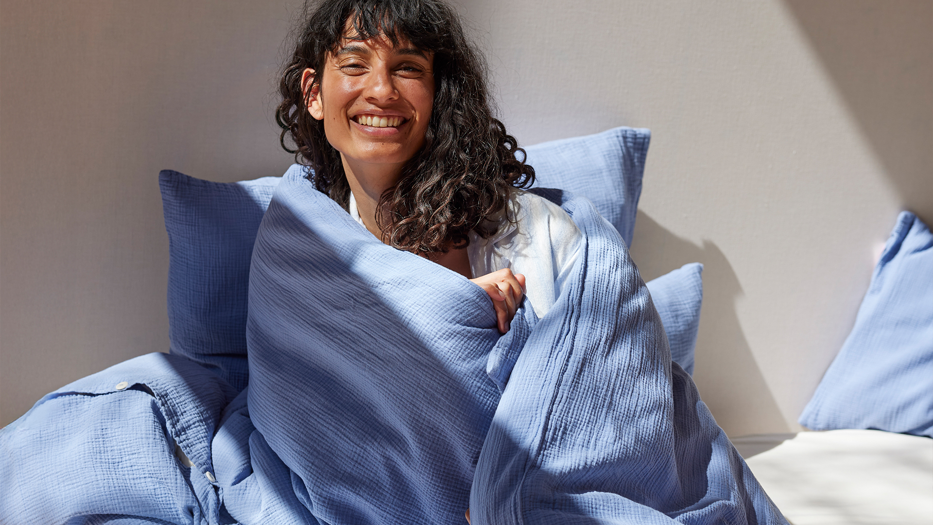 Dunkelhaarige Frau sitzt lächelnd auf einem Bett mit Bettwäsche Vivien in blau