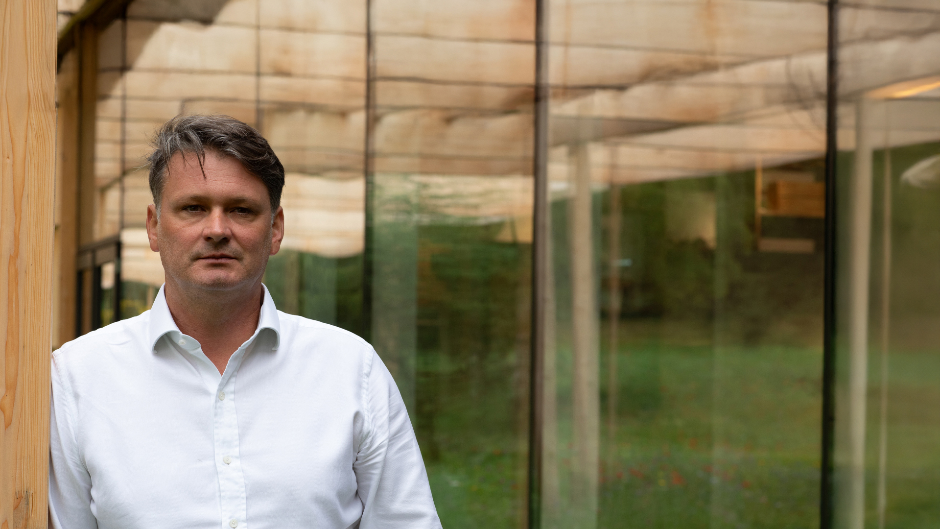 Mann im weißen Hemd steht vor Glasfassade und grüner Umgebung
