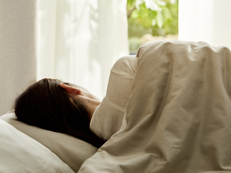 Frau in einem Bett mit ökologischen Schlaftextilien vor einem Fenster mit weißen Vorhängen