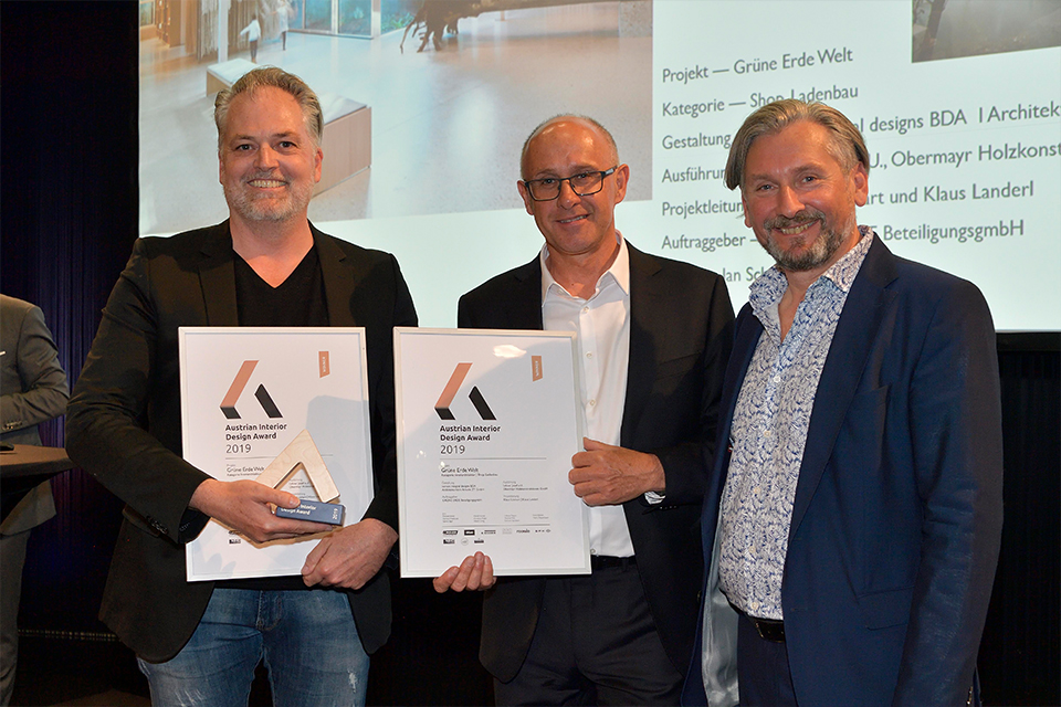 Ein Mann steht neben zwei Männern mit Urkunden bei einer Preisverleihung