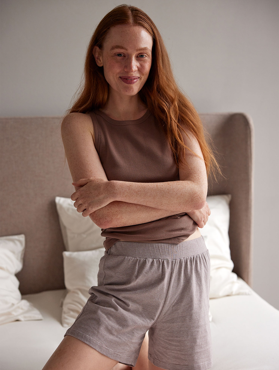 Frau trägt braunes Schlaftop und Shorts und kniet auf einem Bett