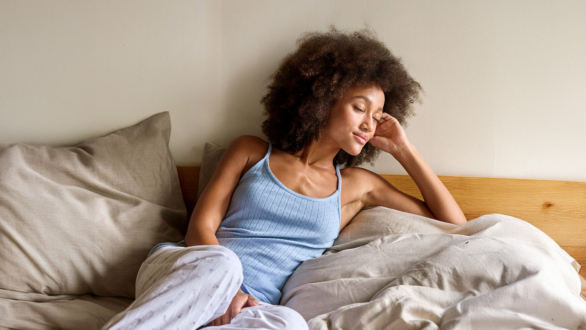 Eine dunkelhaarige Frau mit hellblauem Trägertop in einem Bett mit heller Bettwäsche