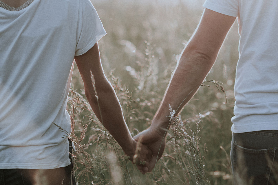 Zwei Personen halten Hände im hohen Gras bei warmem Licht