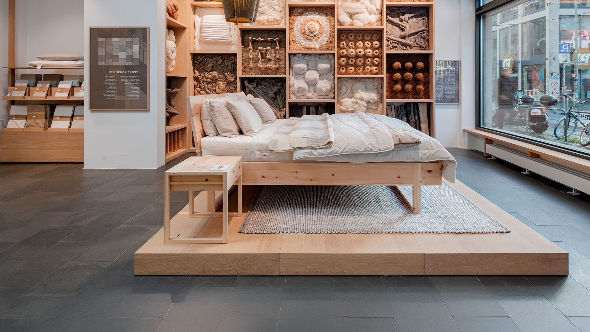 Holzbett auf einem Holzpodest und Naturmaterialien im Hintergrund zur Ansicht im Store