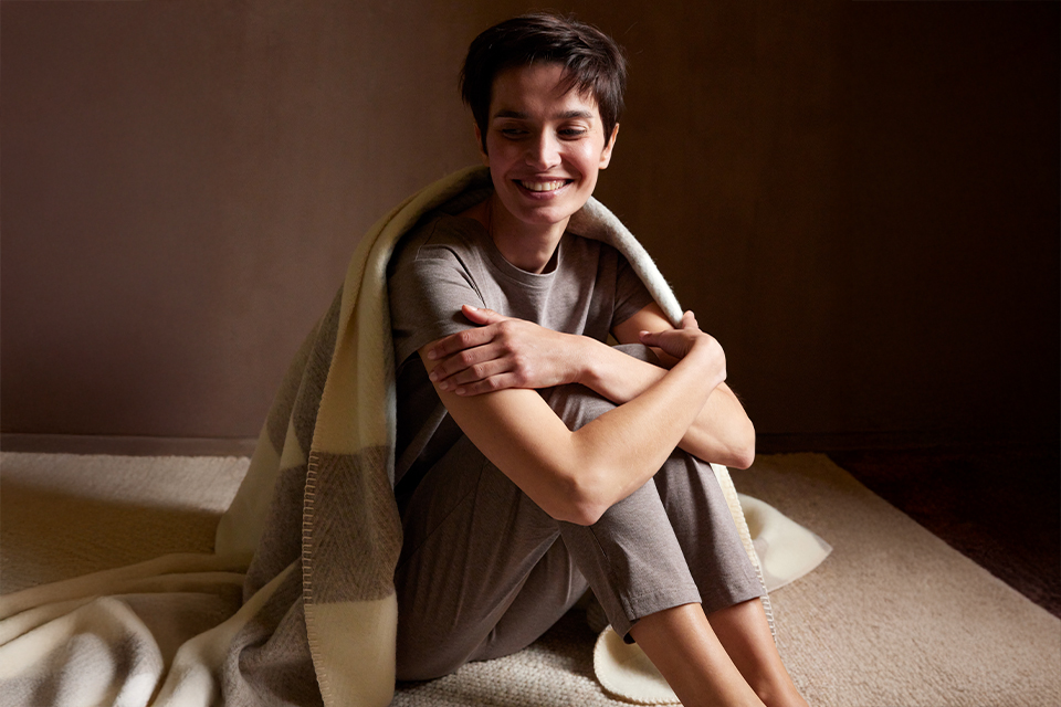 Junge Person sitzt entspannt eingehüllt in Decke auf Teppich