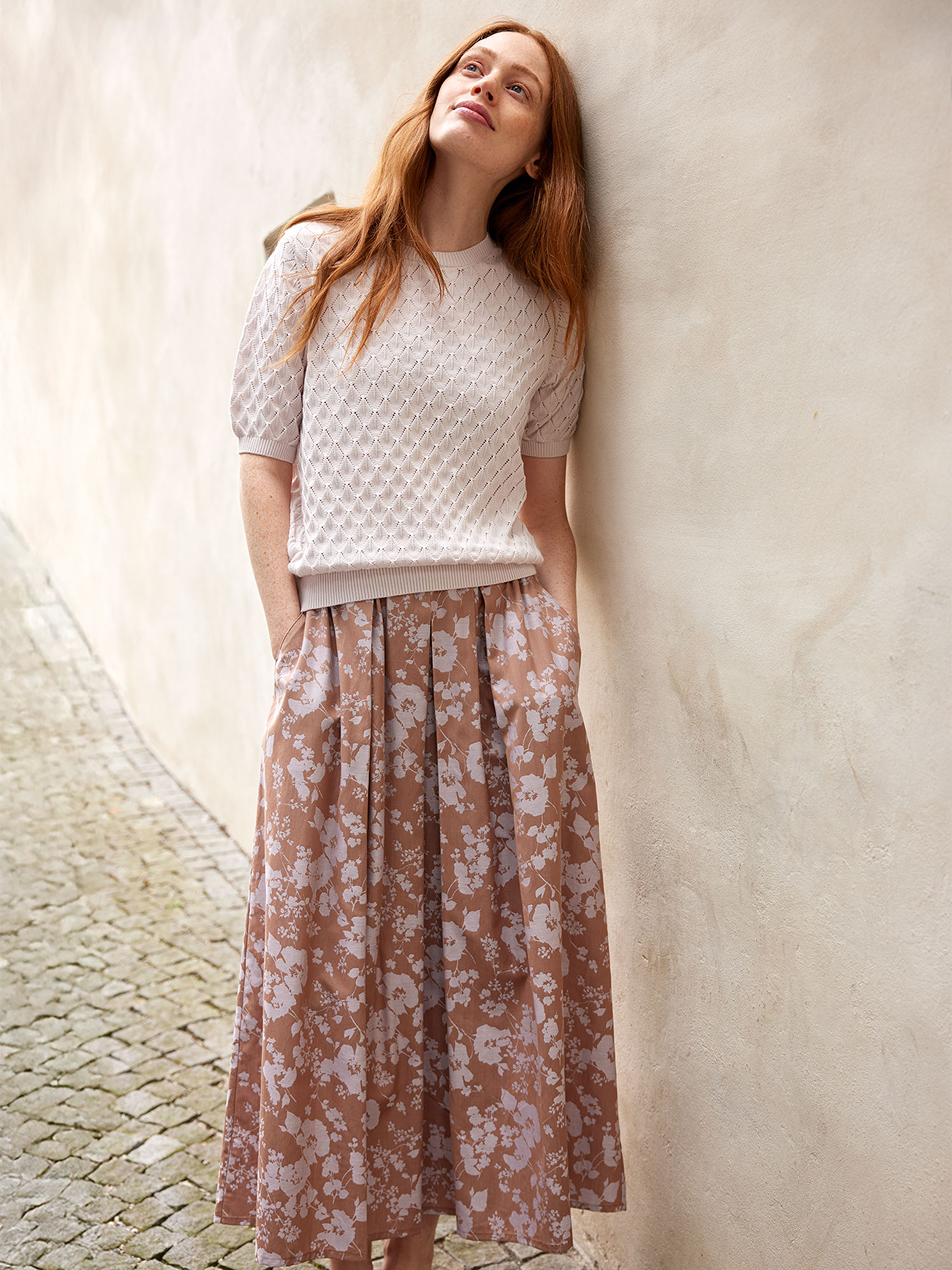 Frau trägt gemusterten Rock und helles Oberteil und lehnt an Wand