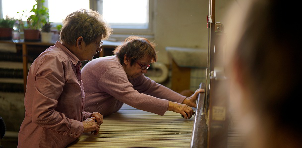 Zwei Frauen bedienen in Handarbeit einen alten Teppich-Webstuhl. 