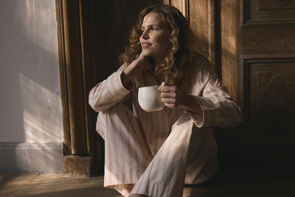 Eine Frau in gestreiftem Pyjama sitzt vor einer Türe am Boden und hält eine Tasse