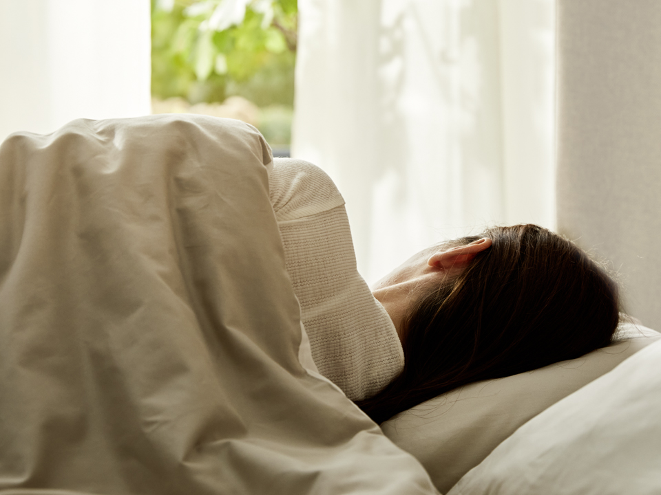 Eine Frau schläft in einem Bett das vor einem Fenster mit weißen Vorhängen steht