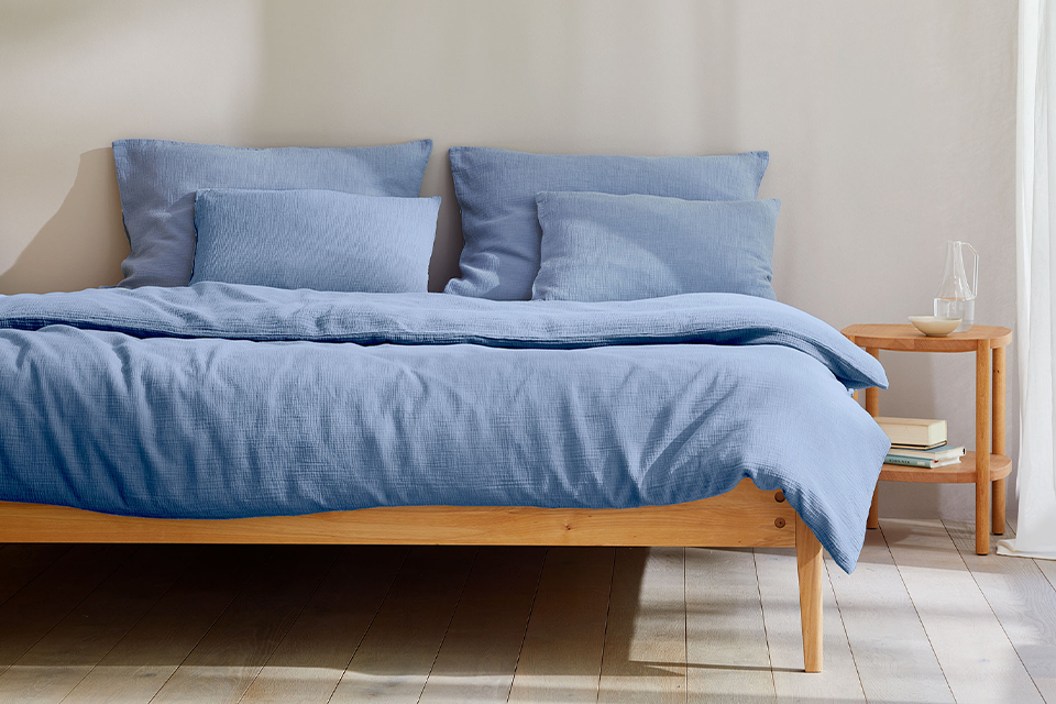 Massivholzbett mit blauer Bettwäsche steht vor einer hellgrauen Wand