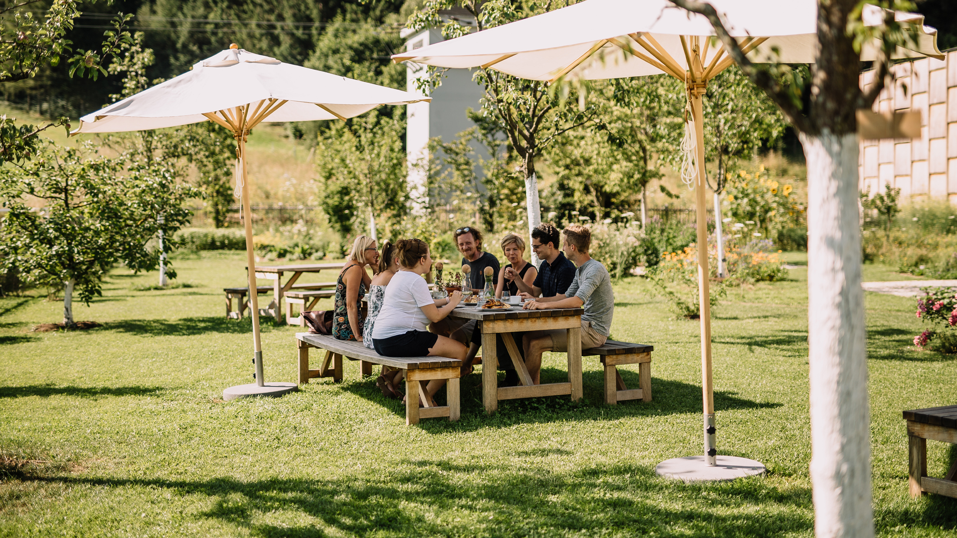 Menschen sitzen gemeinsam im Garten unter Sonnenschirmen am Tisch