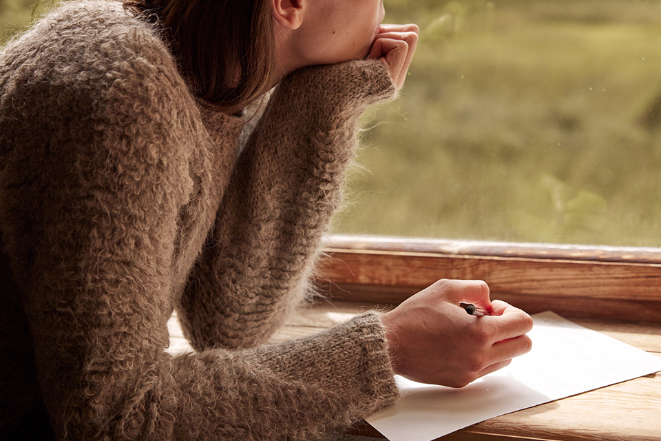 Person sitzt am Fenster und schreibt nachdenklich in Notizbuch