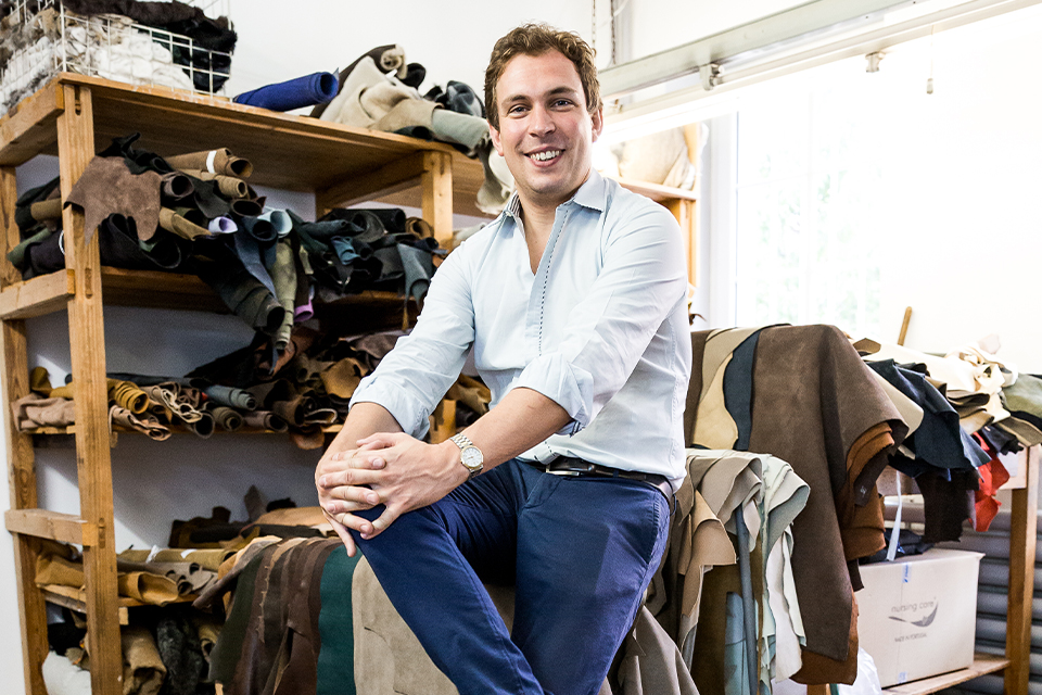 Mann sitzt lächelnd zwischen Stoffrollen und Leder in Werkstatt