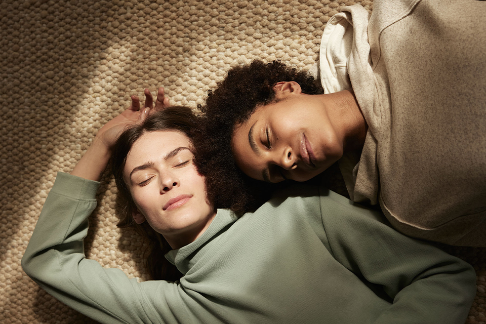 Zwei Frauen in Pullovern liegen auf einem naturfarbenen Teppich