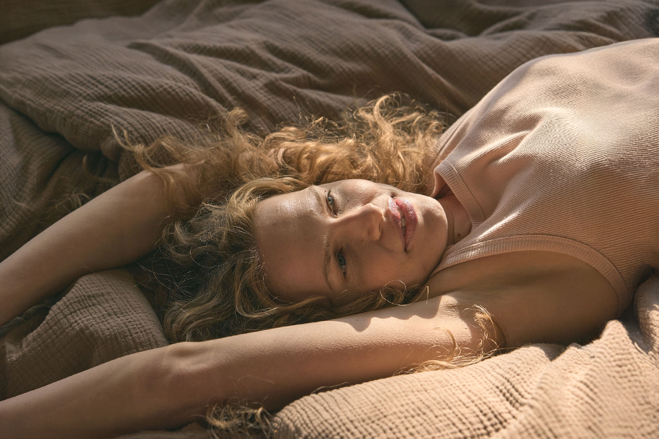 Eine blonde Frau liegt mit ausgestreckten Armen auf einem Bett mit Bettwäsche Vivien in der Farbe nougat