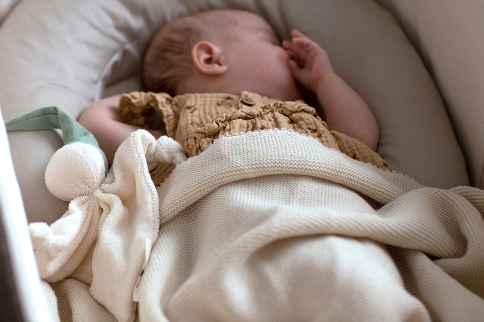 Ein schlafendes Baby in einer Wiege mit heller Kuscheldecke und Schmusewichtel
