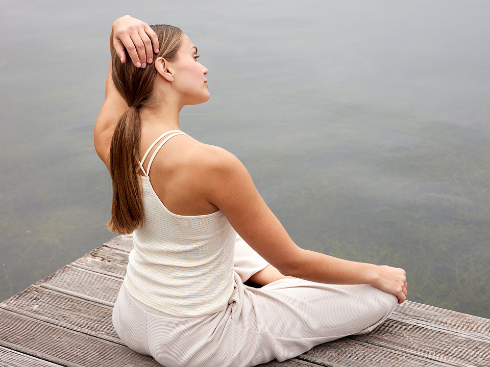 Frau sitzt am Wasser und trägt helles Trägertop und Yogahose