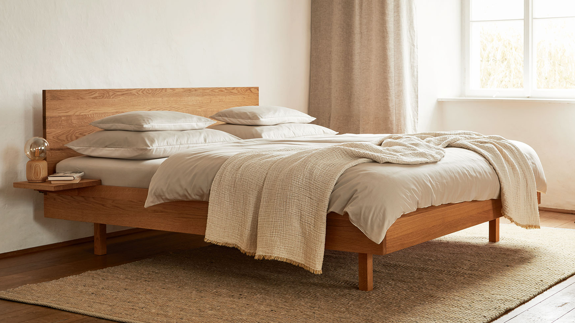 Hochwertiges Bett Tesoro mit heller Bettwäsche und Wohndecke vor einem großen Fenster