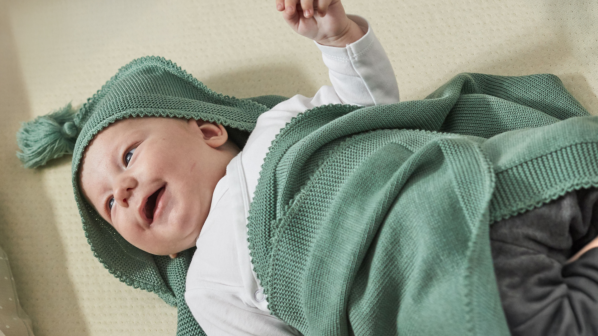 Ein Baby in einer blauen Decke eingewickelt liegt auf einer Matratze und lacht