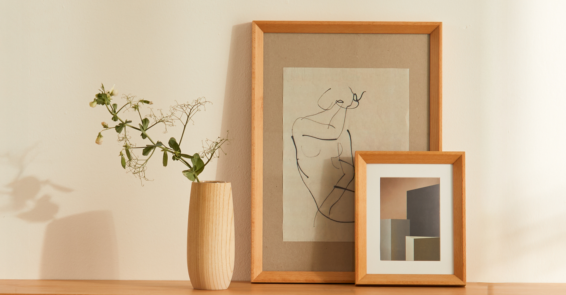 Ein großer Bilderrahmen lehnt an der Wand. Ein kleiner Bilderrahmen lehnt daran. Daneben steht eine Vase mit einer Blume.