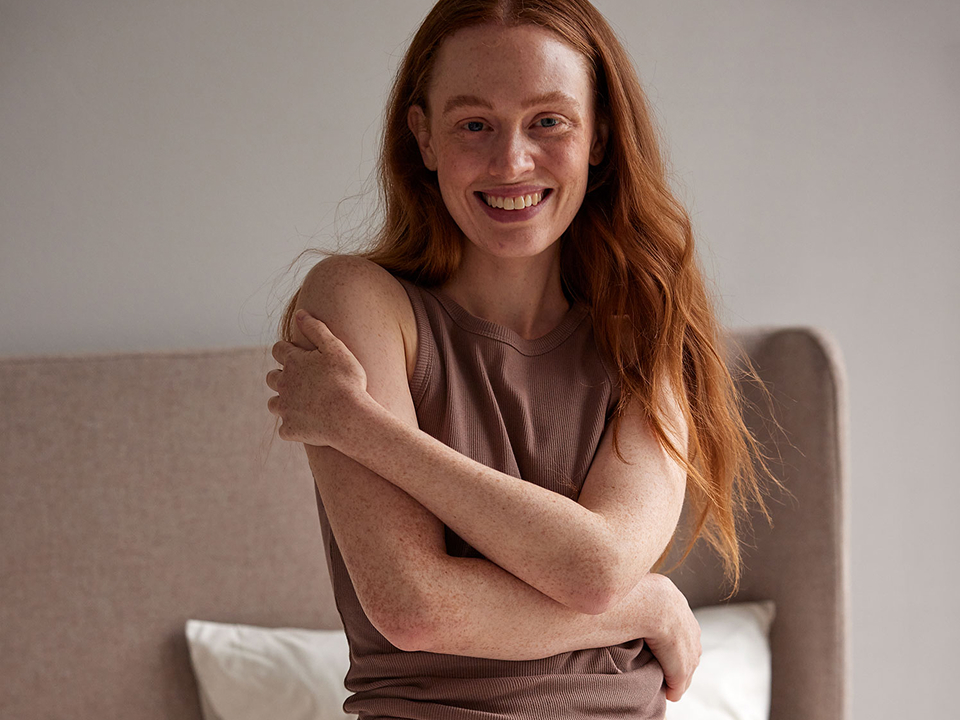 Frau trägt braunes Schlaftop und sitzt entspannt auf einem Bett