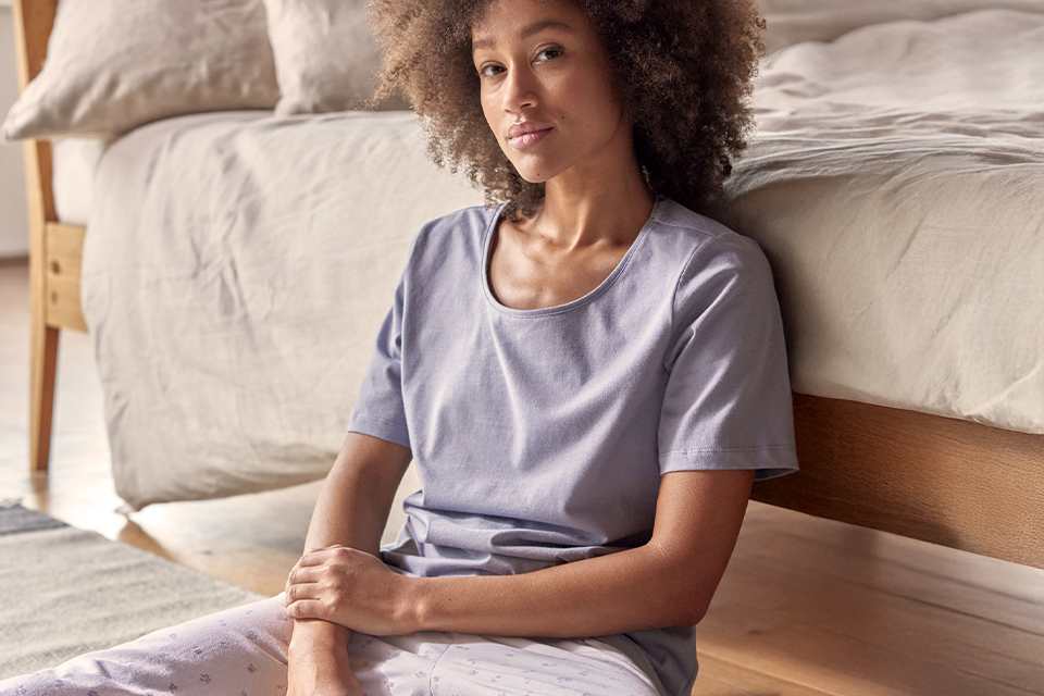 Frau trägt violettes Schlafshirt und sitzt entspannt auf dem Boden neben einem Bett