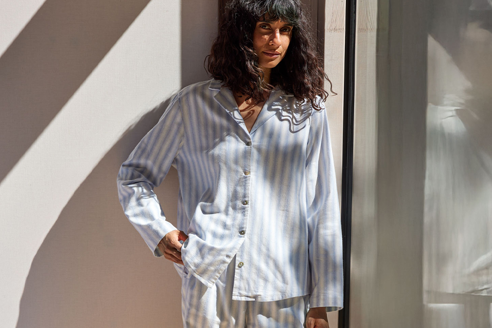 Eine dunkelhaarige Frau trägt einen blau weiß gestreiften Pyjama und steht vor einer hellen Wand